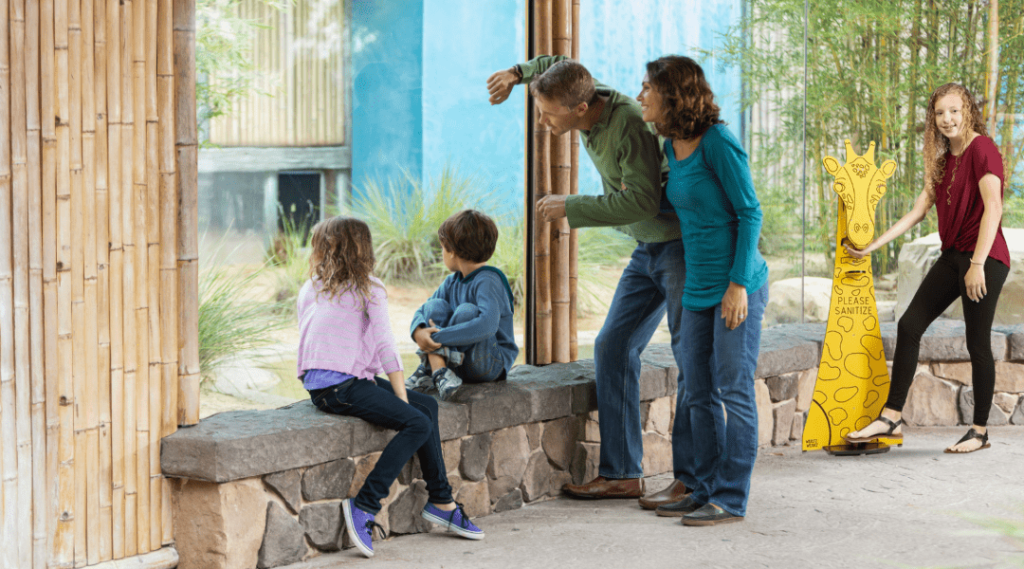 Family looking at zoo animals and nearby is a girl using an animal hand sanitizing station. It is carved into a giraffe and made out of King ColorCore.